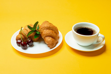 French croissants, cup of coffee and cherries on yellow background close up	