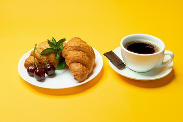 French croissants, cup of coffee, dark chocolate and cherries on yellow background close up	