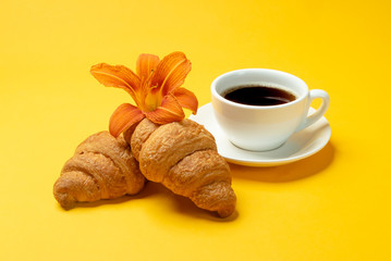 French croissants, cup of coffee and lily flower on yellow background close up