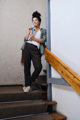 Young joyful woman with dark curly hair in khaki shirt and pants standing on stairs in university holding textbooks in hand and happily looking aside