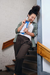 Young attractive girl with dark curly hair in khaki shirt and pants standing on stairs in university holding textbooks in hand and thoughtfully looking down