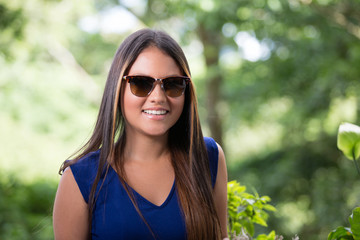 Closeup portrait of beautiful lady at window with nature as background. She is smiling to the camera. She has long and smooth hair, bright eyes, nice smile and sun tanned skin. She is wearing sunglass