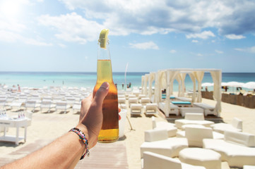 Male hand holding a bottle of beer against a sunny sky and crystal clear sea. In the background a beautiful beach resort. Vacation concept.
