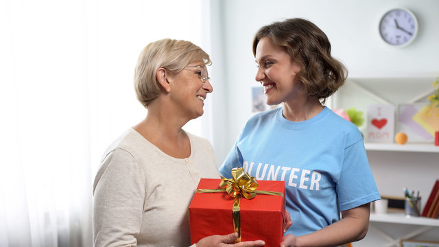 Smiling lady in volunteer t-shirt giving gift box to aged lady, holiday charity