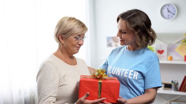 Happy woman in volunteer t-shirt giving gift box to aged lady, holiday charity