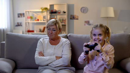 Cheerful schoolgirl playing video game with joystick alone, ignoring granny