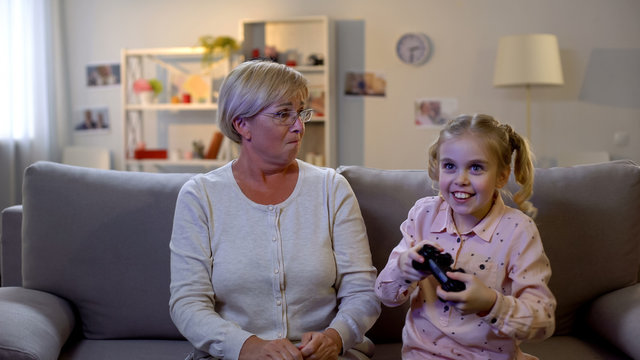 Happy girl playing video game, upset granny enviously looking at granddaughter