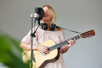 Beautiful blonde singer girl in headphones with a guitar in home recording studio sings a song into a microphone