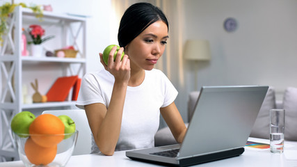 Female freelancer in white t-shirt working online project holding fresh apple