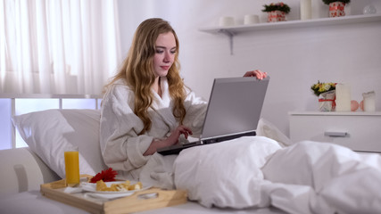 Beautiful female chatting with friend on laptop, breakfast in bed, lazy morning