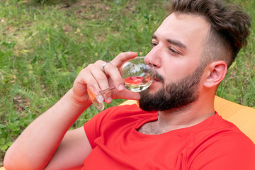 Brutal guy with a beard in a red T-shirt drinks wine. Caucasian traveler on vacation in the woods uses alcohol. Close-up. Portrait of a handsome young man.