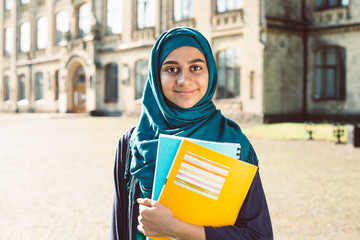 Smiling muslim young female student holding books standing near college. Happy Arabian girl in...
