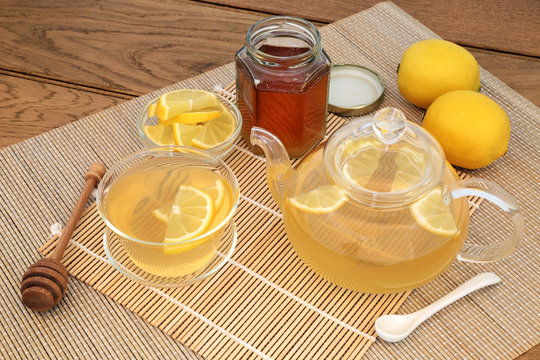 Honey and lemon hot drink to soothe cold, flu and sore throats with fresh lemon fruit, glass teapot and cup, with jar of honey on bamboo and oak background.