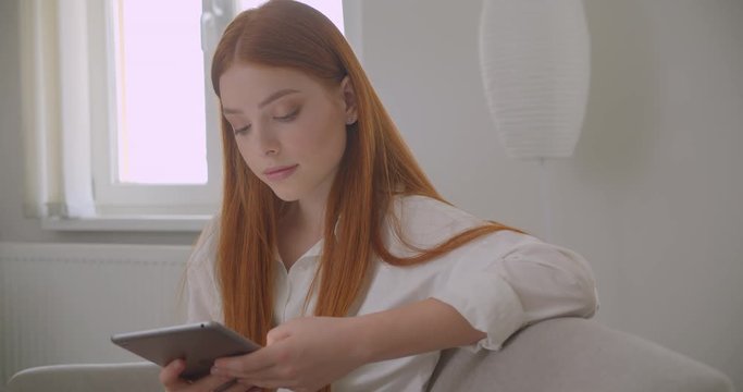 Closeup portrait of young pretty redhead female browsing the tablet and smiling cheerfully sitting on the couch in a cozy apartment