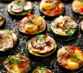 Zucchini pizza bites from zucchini slices with the addition of brown mushrooms, mushroom chanterelles, mozzarella cheese, salami sausage, cherry tomatoes and herbs on a black background, close-up. 