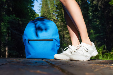 Female legs in white sneakers next to a blue small backpack on a wooden path in the forest. In the background are trees. The concept of hiking, outdoor activities. Place for text.