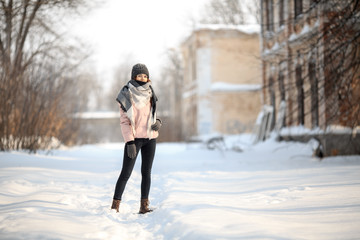 Young cute white caucasian girl in pink down jacket, cap, warm scarf and gloves stands on the street in winter.