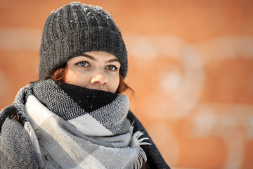 A young cute white girl in a hat and warm scarf is standing in the street opposite red background in winter. Close up.