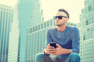 Young American Man traveling in New York City, wearing gray, long sleeve T shirt, sunglasses, sitting in front of business district with high buildings, texting on cell phone, looking, thinking..