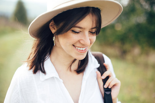 Portrait of happy hipster girl with backpack traveling on top of sunny mountain. Stylish woman in hat smiling and enjoying hiking in mountains. Wanderlust and travel. Atmospheric moment