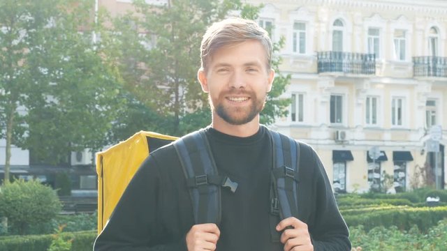 Portrait of a smiling young courier with a yellow bag in the city. Bearded guy with a backpack during the dawn. Delivery service worker close up 4k