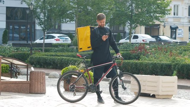 A courier with a yellow bag and a bicycle is looking for the client's address in the phone. Young delivery man with a sports bike 4k