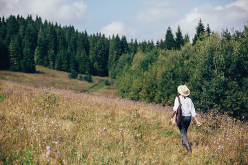 Fototapeta na wymiar Hipster girl with backpack traveling in sunny mountains, walking in sunny wildflower meadow. Stylish woman in hat exploring and hiking on top of mountain. Wanderlust and travel concept. Space