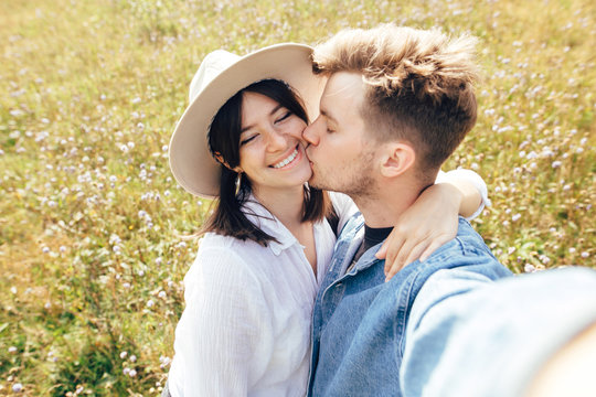 Happy hipster couple making selfie and kissing in sunny mountains. Stylish young family taking photo in travel and enjoying hiking on top of mountain in wildflower meadow. Summer vacation