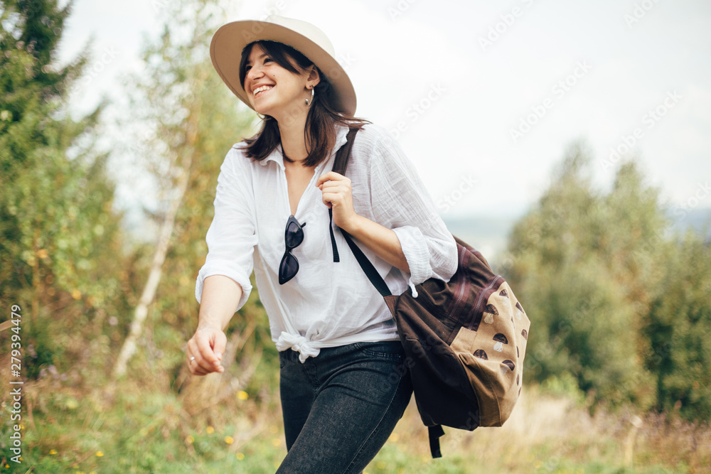 Wall mural hipster girl with backpack traveling on top of sunny mountain, walking on hills with woods. stylish 