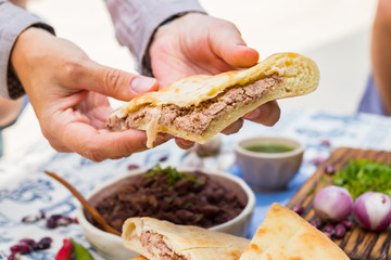 Lobiani pie - Georgian cuisine traditional food stuffed with red kidney or white beans (lobio) inside on wooden board and Lurji Supra tablecloth. Woman hand holds, take lobiani pie piece.