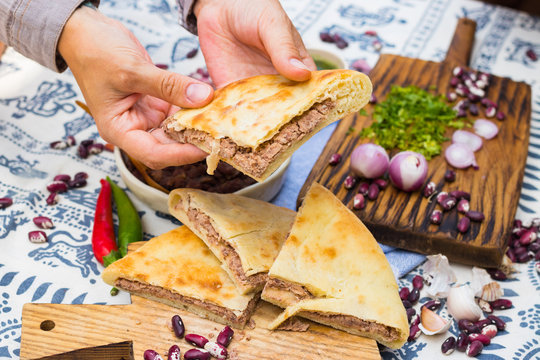Lobiani pie - Georgian cuisine traditional food stuffed with red kidney or white beans (lobio) inside on wooden board and Lurji Supra tablecloth. Woman hand holds, take lobiani pie piece.