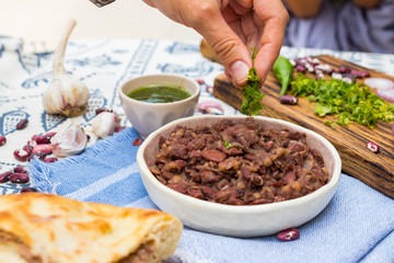 Lobio - Georgian cuisine traditional food with cooked red kidney beans, cilantro coriander and spices on Lurdji Supra tablecloth. With tkemali sauce and lobiani pie. Hand holds lobio.