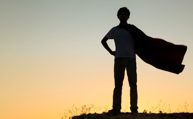 Boy playing superheroes on the sky background, teenage superhero in a red cloak on hill