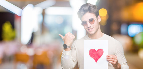 Young handsome man holding card with red heart over isolated background pointing and showing with thumb up to the side with happy face smiling