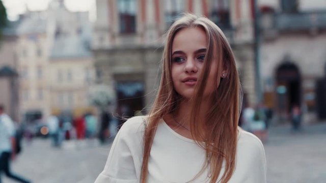 Fancy young blue-eyed blonde woman temptingly walking down the city center, attractively smiling straight to camera. Confidence, female power, natural beauty. Female portrait, slow motion