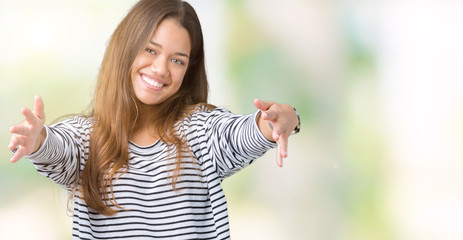 Young beautiful brunette woman wearing stripes sweater over isolated background looking at the camera smiling with open arms for hug. Cheerful expression embracing happiness.