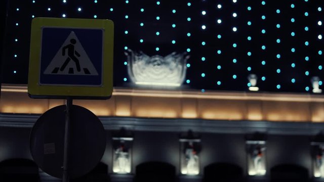 Close-up view of pedestrian crossing sign against the modern shopping center with the blinking night illumination and plaster statues along the wall at night. Stock footage. Beautiful urban buildings