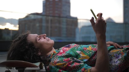 Side view of handsome smiling skater lying with head on skateboard and scrolling social media content on smartphone outdoor. Young man skateboarder laughing while networking on cellphone after riding - Powered by Adobe