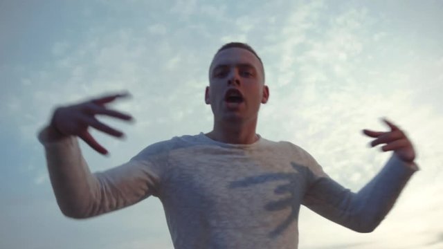 Low angle shot of handsome young Caucasian man rapping and actively gesticulating on cloudy sky background. Music, talent, music video, lyrics. Close up view, male portrait