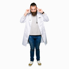 Young blond doctor man with beard wearing medical coat Smiling pulling ears with fingers, funny gesture. Audition problem