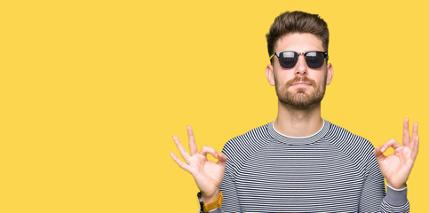 Young handsome man wearing sunglasses relax and smiling with eyes closed doing meditation gesture with fingers. Yoga concept.