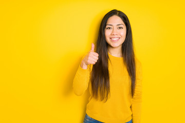 Beautiful brunette woman over yellow isolated background doing happy thumbs up gesture with hand. Approving expression looking at the camera showing success.