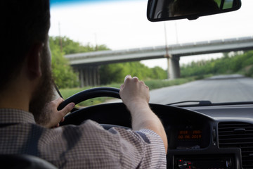 Young bearded man in summer shirt driving a car on highway. Both hands holding a steering wheel.