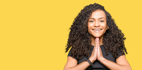 Young beautiful woman with curly hair praying with hands together asking for forgiveness smiling...