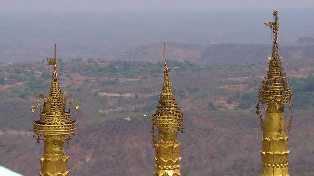 A close up of three golden sculptures on the top of temple and a valley covered with trees at the far background