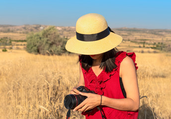 Woman in straw hat and red short holds a camera. Woman preparing to take photo in a yellow field