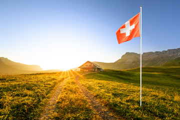 swiss chalet or farm in mountain landscape at sunrise with sun star and swiss flag waving in the foreground 