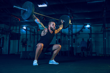 Young healthy man athlete doing exercise with the barbell in the gym. Single male model training hard and practicing in squats. Concept of healthy lifestyle, sport, fitness, bodybuilding, crossfit.