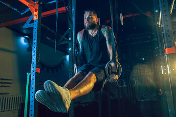 Young healthy man athlete doing exercise with the rings in the gym. Single male model training hard and practicing in pull-ups. Concept of healthy lifestyle, sport, fitness, bodybuilding, crossfit.