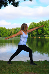 Long-haired blonde girl doing stretching in a park near a lake. Trees and vegetation in bokeh background. Woman doing fitness exercises at the water's edge.